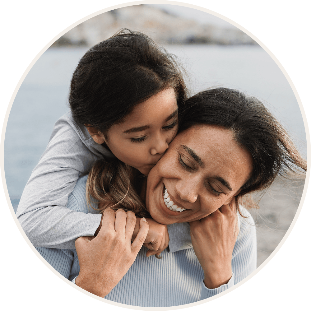 A joyful mother and daughter embrace near a body of water, with the daughter hugging and kissing her mother from behind