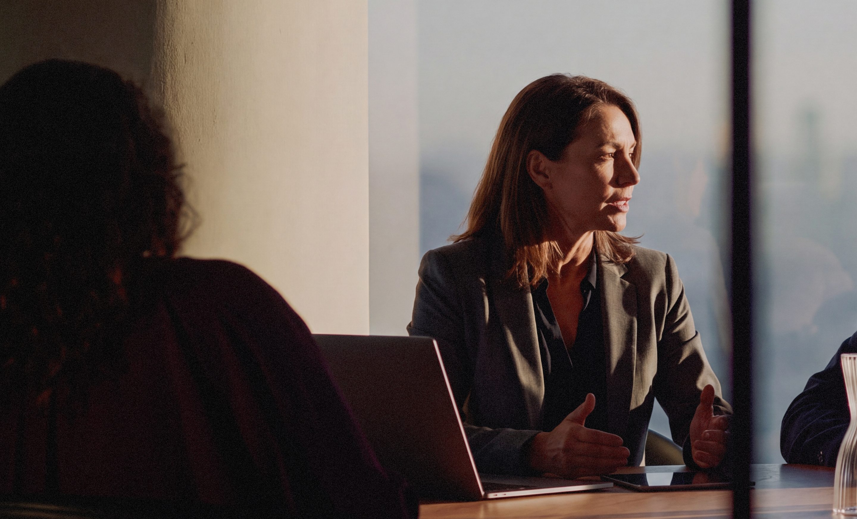 A woman speaking at a meeting table, a city window behind her