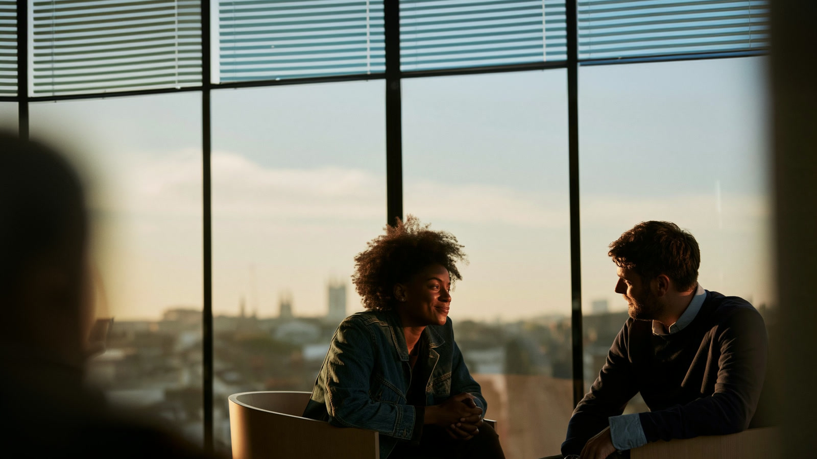 Two coworkers talking by tall office windows at dusk